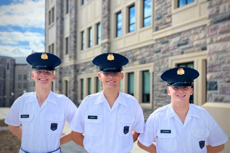Cadets in white uniform shirts stand smiling with Hokie Stone and blue skies in the background.