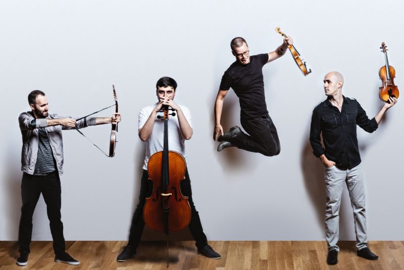 The members of Invoke, four white men wearing casual cloths, playfully pose in front of a light grey background. One member pulls back the strap on his mandolin like a bow and arrow and one jumps into the air.