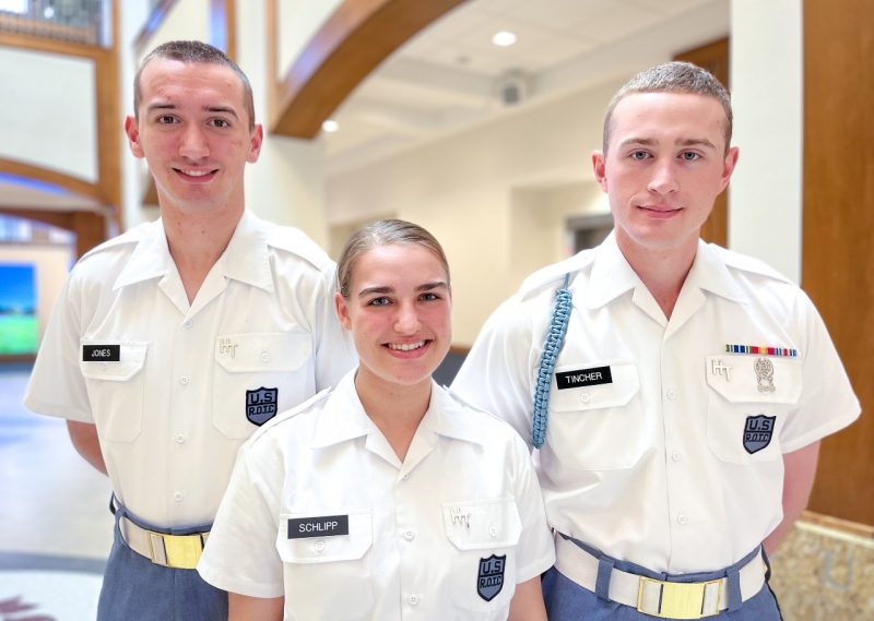 Cadets in white uniform shirts stand smiling in the atrium of the Corps Leadership and Military Science Building.