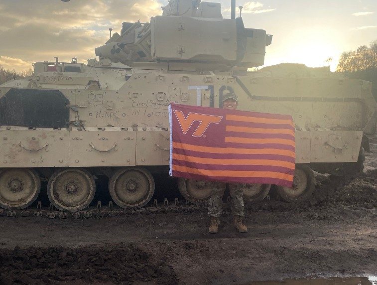 Kim smiles while holding a striped Virginia Tech flag. She is wearing her camouflage uniform in front of a large military tank and the sun is low in the sky. 