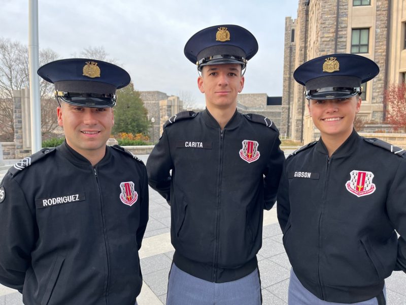 The three cadets stand in their black uniform jackets and military hats near the flagpole on Upper Quad. Hokie stone buildings are in the background and the cadets are smiling. 