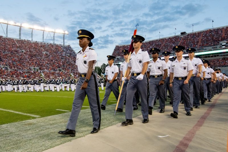 Cadet Heron marches in front of a group of cadets on a football field with crowded stands and stadium lights in the background. 