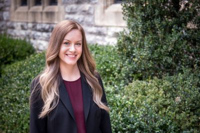 woman in blazer poses in front of hedge and stone wall