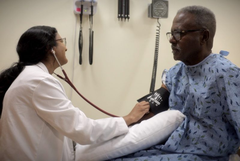 African American man wearing patient gown having is blood pressure taken by a medical student wearing a white coat.