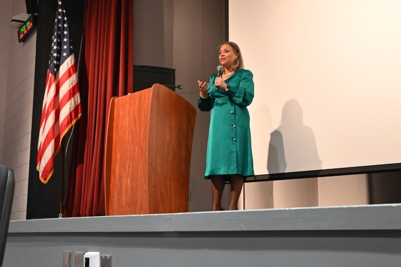 Janice Underwood in a jade satin dress speaks from a podium while standing in a warm-toned spotlight.