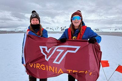 Two people hold a flag with the Virginia Tech logo on snowy ground, with tall mountains behind them. 
