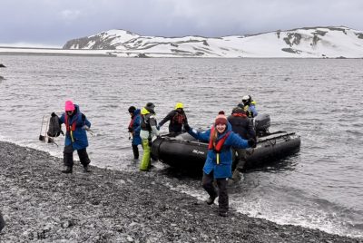 An inflatable zodiac boat is in shallow water as people disembark onto a rocky beach, with snow-covered hills in the background.