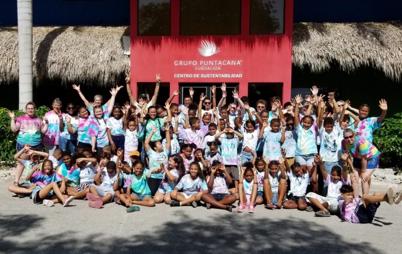 A large group of children in tie dyed shirts poses for a photo in front a building, smiling and waving their arms,