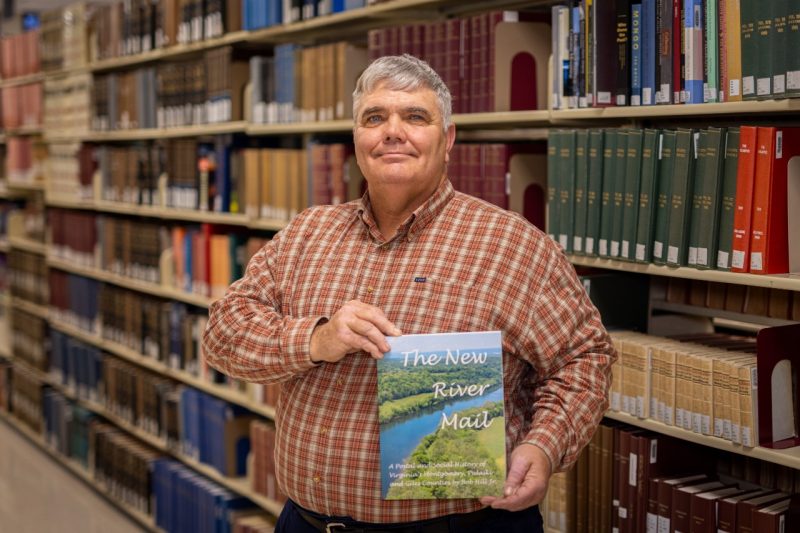 Bob Hill stands among the stacks in Newman Library with his book.