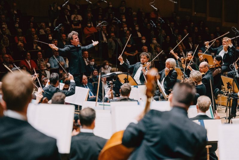 Conductor Jakub Hrůša, a middle aged white man with sandy brown hair, extends his arms out wide as he conducts the Bamberg Symphony during a live performance. All the string players watch him and their bows are raised in the air away from their instruments.