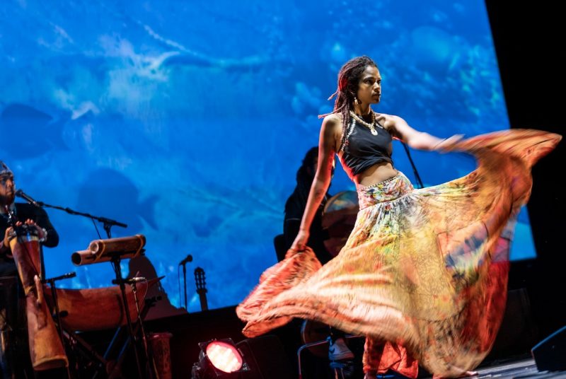 Indigenous woman wears a long flowing orange skirt, which is blurred as she twirls it on stage.