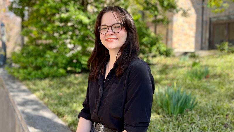 Woman with long dark hair and glasses, wearing a black shirt, sits on a wall with greenery behind her.