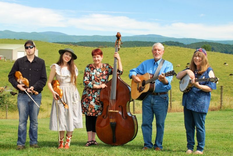 The members of the Whitetop Mountain Band hold their instruments and stand in a green field, mountain ranges visible in the distance behind them. From left, a young white man holding a fiddle in a black button down shirt and jeans, a young white woman with long brown hair holding a fiddle and wearing a light pink dress, a middle aged white woman with short red-brown hair and holding a double bass, an older white man with white hair holding an acoustic guitar and wearing a blue button down shirt and jeans, and a middle aged white woman with medium length brown hair holding a banjo and wearing a denim shirt and denim pants.
