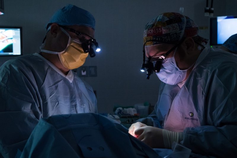 Dark room, two indivduals in surgical attire looking down at a patient.