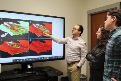 A person gestures to a map projected on a screen, as two others look on. 