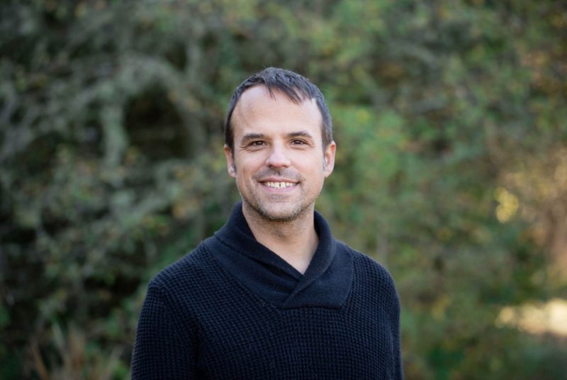 Moss Arts Center Guest Curator Andre Bouchard, a young Indigenous man with short light brown hair and dark brown eyes. He hears a cowl neck black sweater and smiles towards the camera in front of greenery outside.