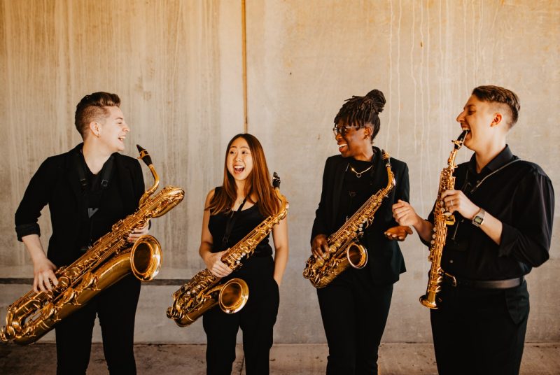 The Medusa Saxophone Quartet, made up of four musicians, all dressed in black, laughing, holding their instruments.