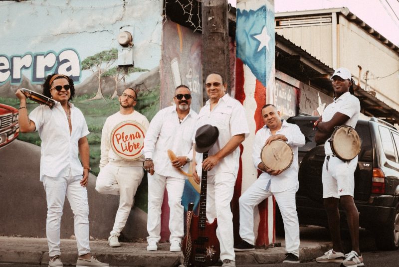 The members of Plena Libre, six Hispanic men, wear all white and hold their instruments on a city street.