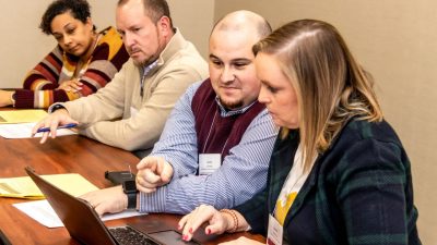 Four people sit at a wooden table. One man points at a laptop screen.