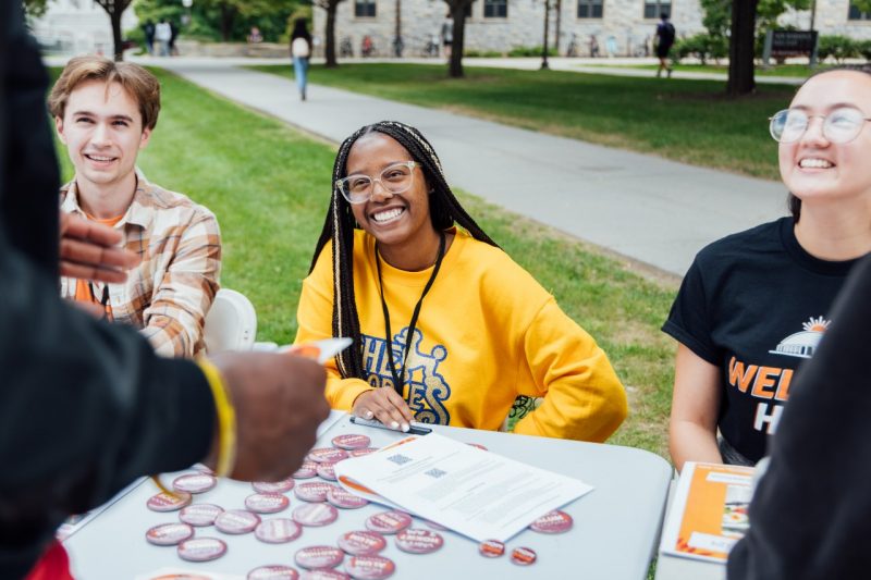 Three students smile and laugh with someone standing in front of their table.