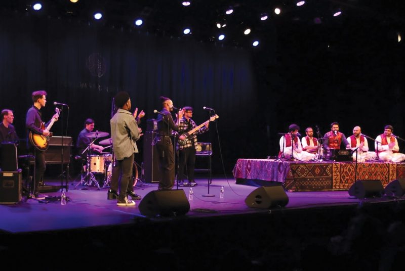 Members of Riyaaz Qawwali and the Harlem Gospel Travelers perform together on stage under purple light. The Riyaaz Qawwali members sit on a raised platform draped in carpets.