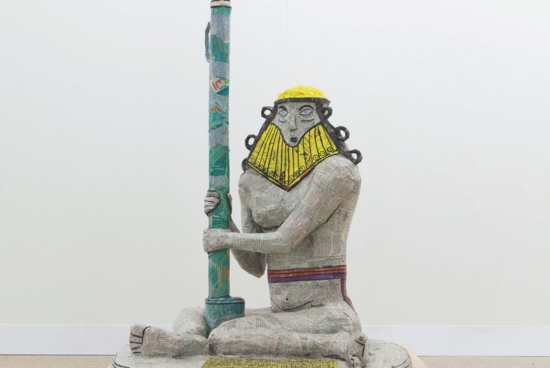 A statue of a male figure with a gold helmet grips a green pole. Michael Rakowitz's work The invisible enemy should not exist – Seated Nude Male Figure, Wearing Belt Around Waist.