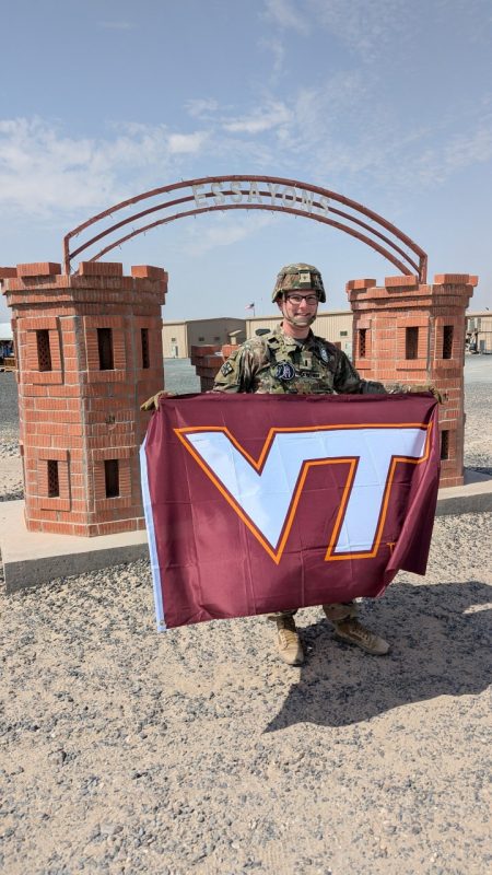 A serviceman in a camouflage uniform and helmet stands in a desert setting holding a Virginia Tech flag. 