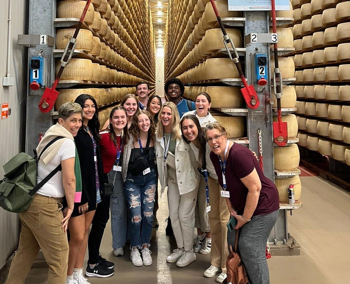 A group of people pose in front of wheels of parmesan, stacked up to the ceiling.