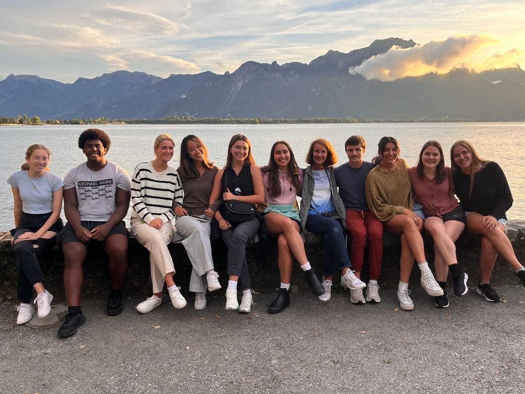 A group of students sits on a wall in front of a Swiss lake.