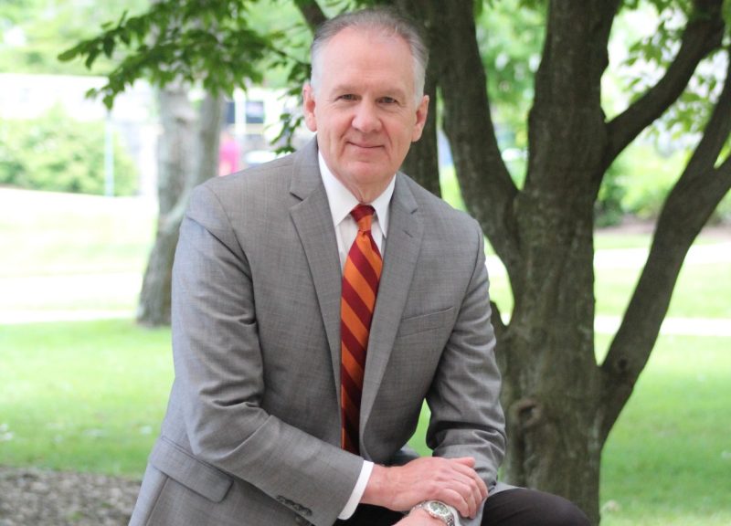 Man with gray hair with an orange and maroon tie in a gray suit jacket.