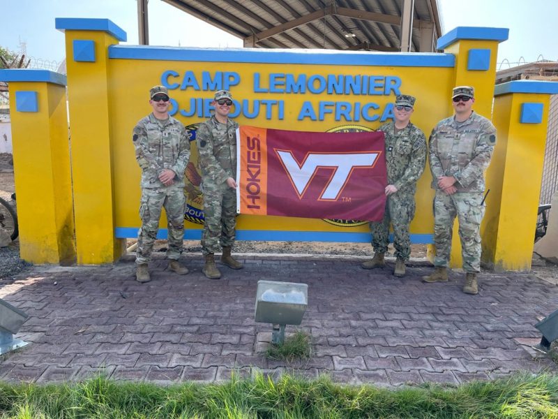 Four service members smile at the camera while holding a Virginia Tech flag. They are in camouflage uniforms standing in front of a yellow sign for Camp Lemonnier. 