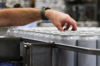 Unlabeled cans on the assembly line. "Brew with purpose" is visible on the cans. 