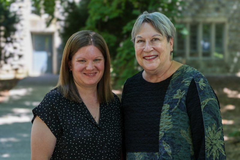 Two individuals pose together on Virginia Tech campus with green foliage and Hokie stone building in the background.