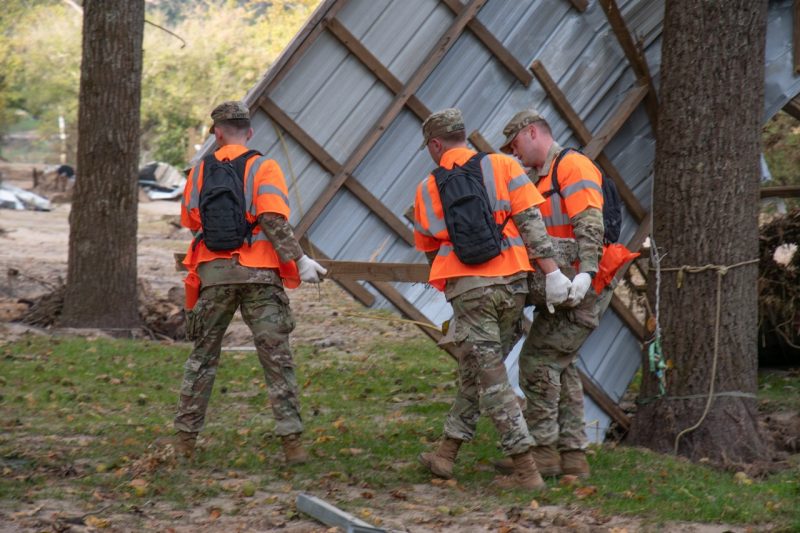 Three cadets carry a post with a cement base past a sideways roof leaning against a tree. 