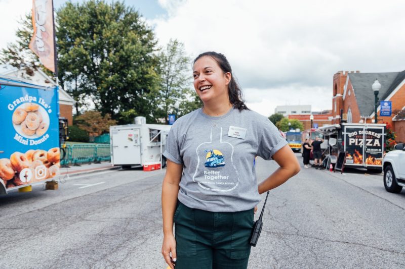 Elli stands in the middle of the street looking off to the left and smiling, surrounded by food trucks on either side and behind her