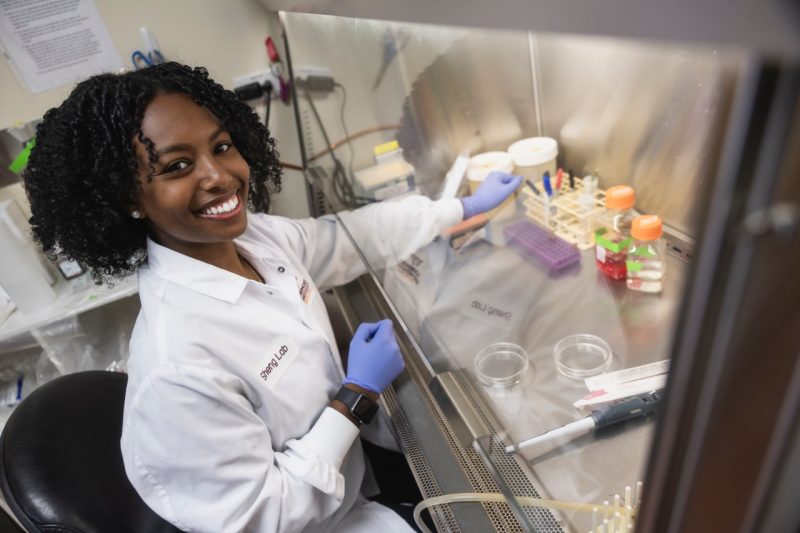 A student in a white coat looks up from lab work and smiles.