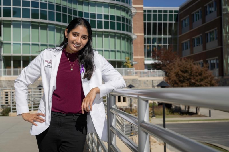 Tara Menon, a medical student, stands against a handrail in front of a glass and brick building.