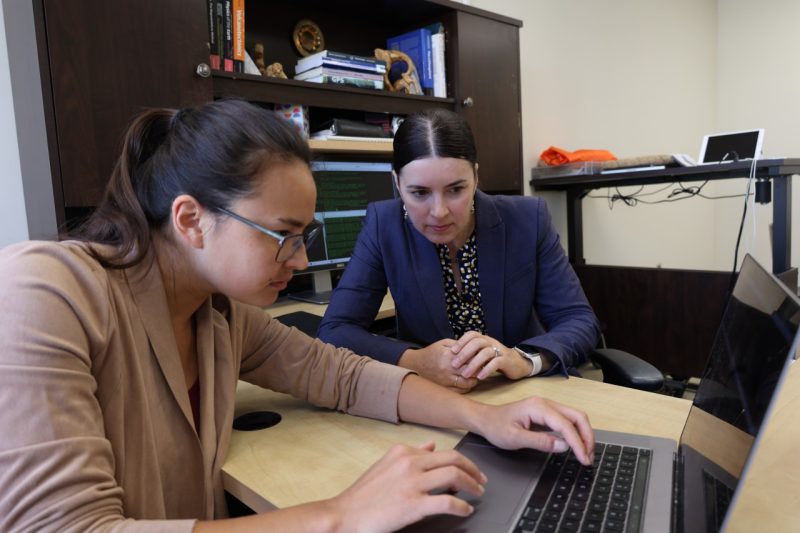 Two women look at laptop screen in office setting.