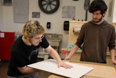 A student presses a piece of paper down onto a block of linoleum with one hand, while the other hand holds up the linoleum from below.