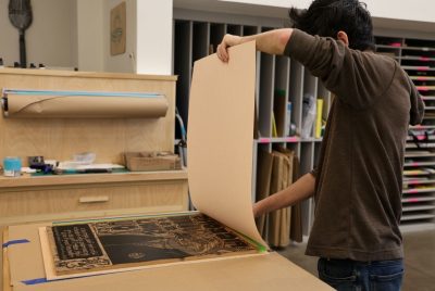 A student holds a large sheet of paper upright while in the process of laying it down on an inked carved wood blood.