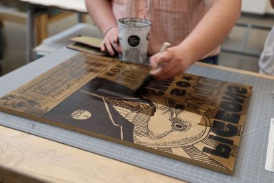 A hand holding a paint brush and a metal can applies a dark liquid to a design on a wood block. 