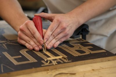 Two hands hold a linoleum cutter being used to carve a design on a flat piece of wood.