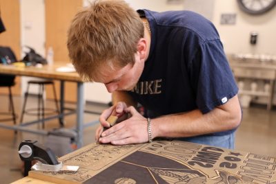 A student hunches over a wood block while carving it with a linoleum cutter.