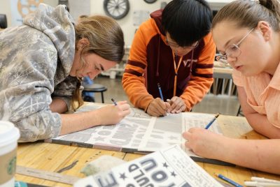 Three students sit around a table, leaning forward and drawing on a piece of paper.