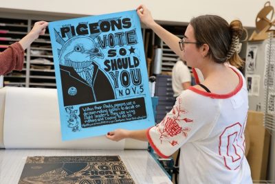 A student holds up a printer poster with a drawing of a pigeon’s head and the words “PIGEONS VOTE SO SHOULD YOU NOV. 5. Within their flocks, pigeons use a decision-making system to decide on flight leaders. They use wing motions to do so.”