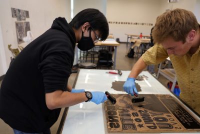 Two students each hold an inker while spreading ink over a carved wood block.