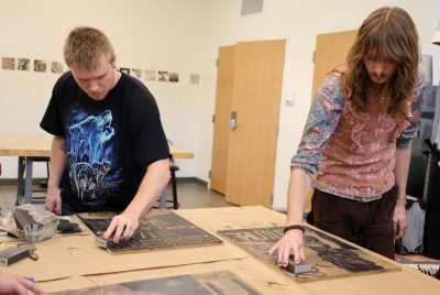 Two students stand beside a wooden table, moving sanding blocks over two carved wood blocks.