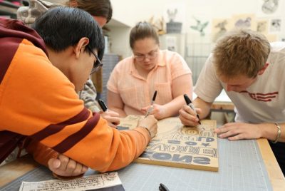 Three students sit at a table, using Sharpies to trace the design on a wood block.