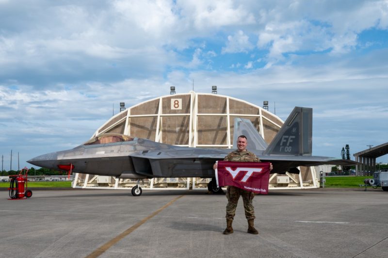A serviceman in a camouflage uniform stands in front of a military fighter jet holding a Virginia Tech flag. 
