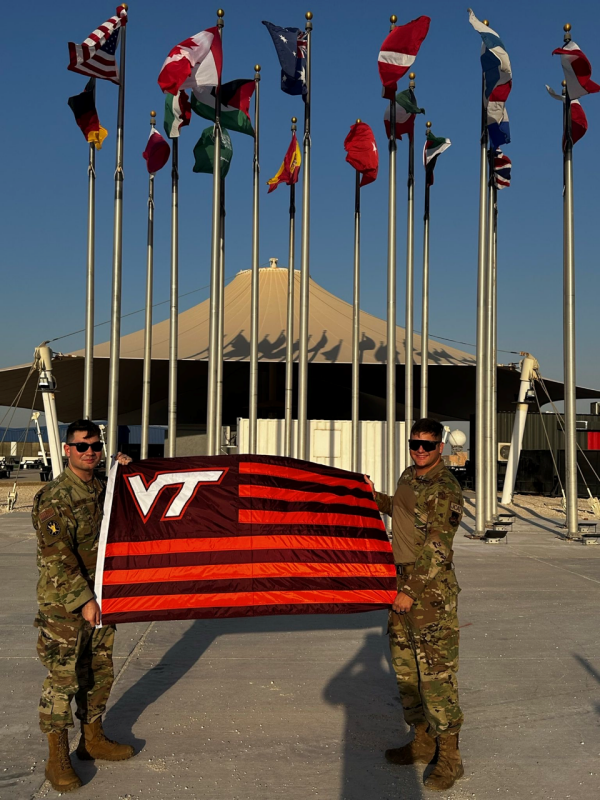 two officers stand in front of a military tent while holding a striped VT flag. They are smiling and wearing camouflage.